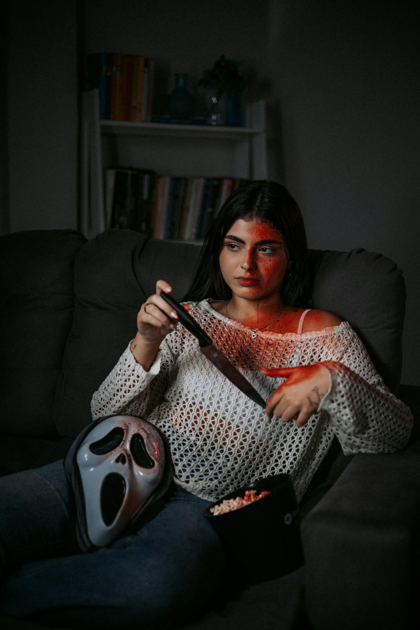 A young woman watches a horror movie on a sofa, holding a knife and popcorn.