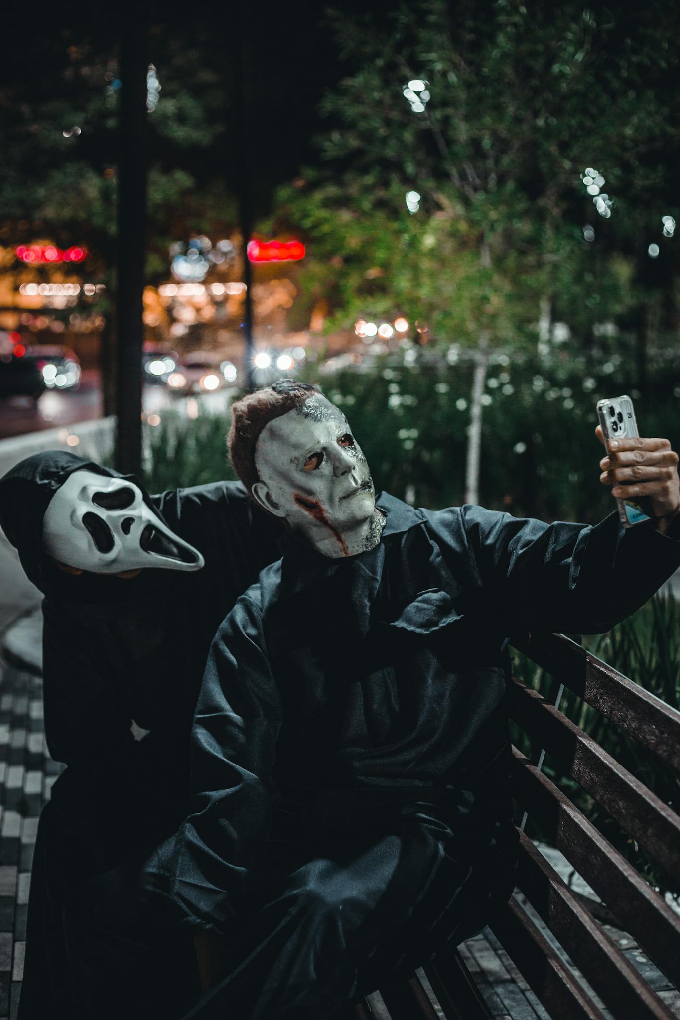 Two masked individuals in a park take a selfie at night, creating a spooky urban scene.
